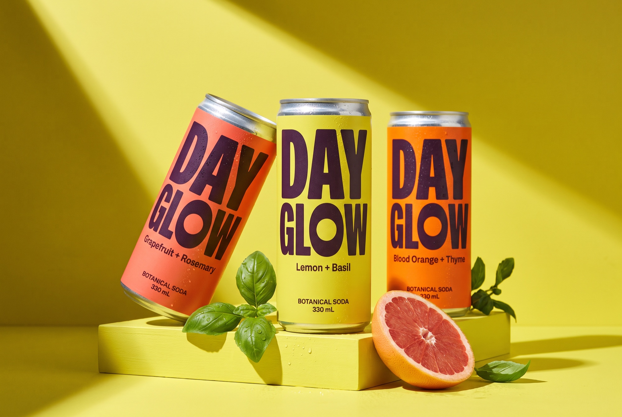 Three DAYGLOW botanical soda cans in yellow, coral and orange, with grapefruit and basil in hard afternoon light.
