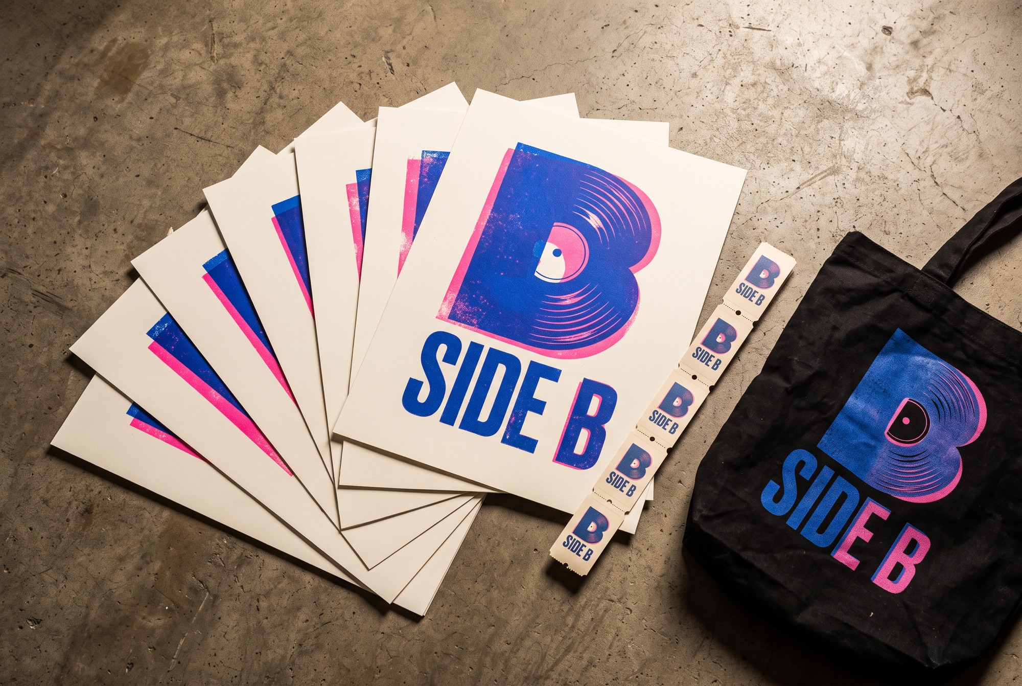 A fanned stack of screen-printed SIDE B posters in cobalt and hot pink with tickets and a black tote.